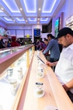 A bright, modern interior features a long display counter with jars of cannabis. People are inspecting and discussing the products. The lighting is a mix of natural light and cool, ambient ceiling lights. Balloons are visible in the background, suggesting a celebratory or event-like atmosphere.