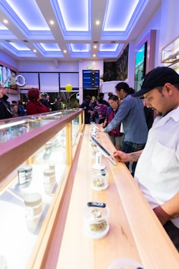 A bright, modern interior features a long display counter with jars of cannabis. People are inspecting and discussing the products. The lighting is a mix of natural light and cool, ambient ceiling lights. Balloons are visible in the background, suggesting a celebratory or event-like atmosphere.