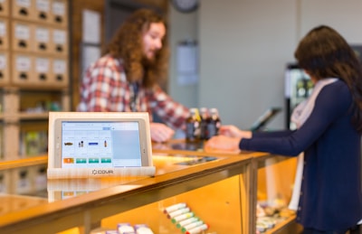 A retail setting with a digital point of sale system on a counter displaying product information. A customer and an employee are engaged in a transaction, with the employee wearing a plaid shirt and the customer holding a bottle. Shelves filled with products are visible in the background.