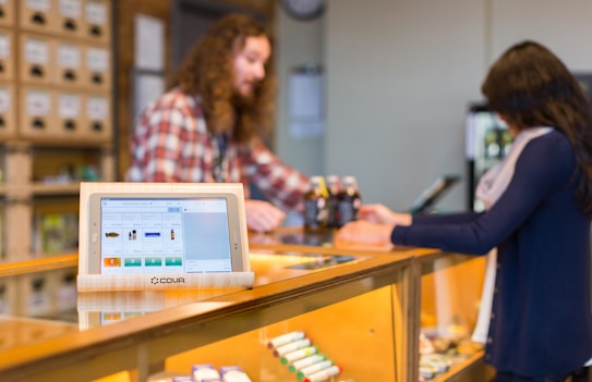 A retail setting with a digital point of sale system on a counter displaying product information. A customer and an employee are engaged in a transaction, with the employee wearing a plaid shirt and the customer holding a bottle. Shelves filled with products are visible in the background.