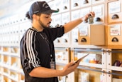 A person wearing a cap and a sports jacket is organizing products in drawers labeled 'Top Shelf' on a wooden shelf, while holding a tablet. The setting appears to be a dispensary or retail space.