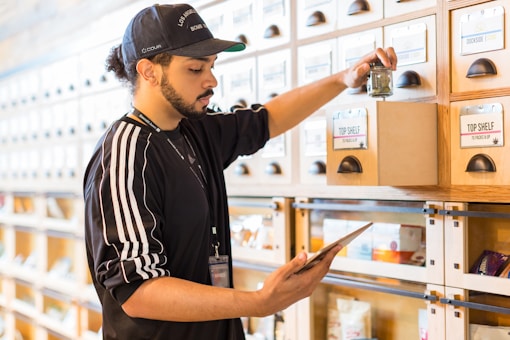 A person wearing a cap and a sports jacket is organizing products in drawers labeled 'Top Shelf' on a wooden shelf, while holding a tablet. The setting appears to be a dispensary or retail space.