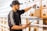A person wearing a cap and a sports jacket is organizing products in drawers labeled 'Top Shelf' on a wooden shelf, while holding a tablet. The setting appears to be a dispensary or retail space.