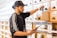 A person wearing a cap and a sports jacket is organizing products in drawers labeled 'Top Shelf' on a wooden shelf, while holding a tablet. The setting appears to be a dispensary or retail space.
