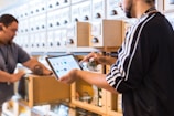 Two people engaged in a workflow process in what appears to be a well-organized storage area. One person is using a tablet device, possibly for inventory management, while holding a small item. The background consists of a series of labeled drawers or cabinets, suggesting an organized system for item storage.