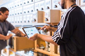 Two people engaged in a workflow process in what appears to be a well-organized storage area. One person is using a tablet device, possibly for inventory management, while holding a small item. The background consists of a series of labeled drawers or cabinets, suggesting an organized system for item storage.