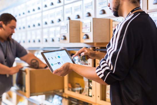 Two people engaged in a workflow process in what appears to be a well-organized storage area. One person is using a tablet device, possibly for inventory management, while holding a small item. The background consists of a series of labeled drawers or cabinets, suggesting an organized system for item storage.