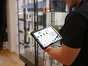 A person holds a tablet displaying a digital catalog in a retail store. The background features glass display cases with various products, and the store interior includes hardwood flooring.