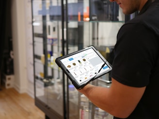 A person holds a tablet displaying a digital catalog in a retail store. The background features glass display cases with various products, and the store interior includes hardwood flooring.