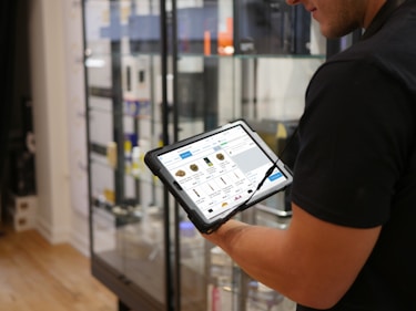A person holds a tablet displaying a digital catalog in a retail store. The background features glass display cases with various products, and the store interior includes hardwood flooring.