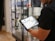 A person holds a tablet displaying a digital catalog in a retail store. The background features glass display cases with various products, and the store interior includes hardwood flooring.