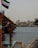 A traditional wooden boat is docked by a body of water with a backdrop of older-style buildings in the distance. The UAE flag is prominently displayed, and a life preserver and fire extinguisher are visible on the boat.