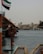 A traditional wooden boat is docked by a body of water with a backdrop of older-style buildings in the distance. The UAE flag is prominently displayed, and a life preserver and fire extinguisher are visible on the boat.