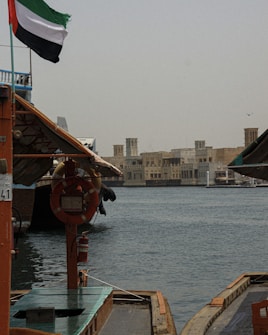 A traditional wooden boat is docked by a body of water with a backdrop of older-style buildings in the distance. The UAE flag is prominently displayed, and a life preserver and fire extinguisher are visible on the boat.