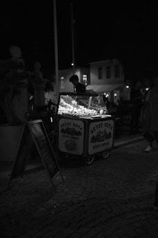 A nighttime scene featuring a street vendor cart illuminated by its own lights. The vendor is standing behind the cart, which displays a variety of items, likely food, given the sign that reads 'SEYFİ USTA MİDYE DOLMA'. The surrounding area is dark, with a few silhouetted figures and dimly visible buildings in the background. The atmosphere is quiet and subdued, typical of a late evening urban setting.