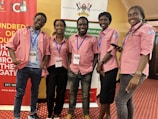 Five individuals are standing together indoors, all wearing matching pink collared shirts with blue lanyards. They are smiling and appear to be at an event hosted by Makerere University, evidenced by the banners in the background. The room has a red carpeted floor and yellow-colored walls.