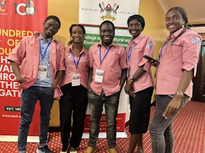 Five individuals are standing together indoors, all wearing matching pink collared shirts with blue lanyards. They are smiling and appear to be at an event hosted by Makerere University, evidenced by the banners in the background. The room has a red carpeted floor and yellow-colored walls.