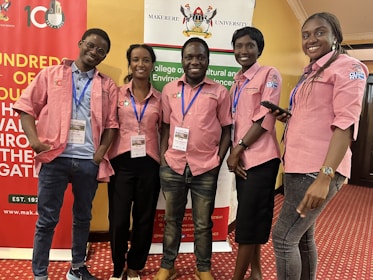 Five individuals are standing together indoors, all wearing matching pink collared shirts with blue lanyards. They are smiling and appear to be at an event hosted by Makerere University, evidenced by the banners in the background. The room has a red carpeted floor and yellow-colored walls.