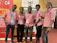 Five individuals are standing together indoors, all wearing matching pink collared shirts with blue lanyards. They are smiling and appear to be at an event hosted by Makerere University, evidenced by the banners in the background. The room has a red carpeted floor and yellow-colored walls.