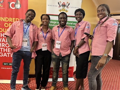 Five individuals are standing together indoors, all wearing matching pink collared shirts with blue lanyards. They are smiling and appear to be at an event hosted by Makerere University, evidenced by the banners in the background. The room has a red carpeted floor and yellow-colored walls.