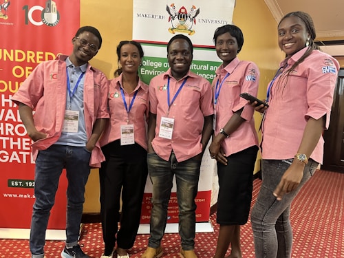 Five individuals are standing together indoors, all wearing matching pink collared shirts with blue lanyards. They are smiling and appear to be at an event hosted by Makerere University, evidenced by the banners in the background. The room has a red carpeted floor and yellow-colored walls.