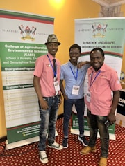 Three individuals are standing in front of two vertical banners related to Makerere University. They are smiling and appear to be at an event or conference. The individuals are wearing casual clothes and have name tags around their necks.
