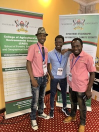 Three individuals are standing in front of two vertical banners related to Makerere University. They are smiling and appear to be at an event or conference. The individuals are wearing casual clothes and have name tags around their necks.