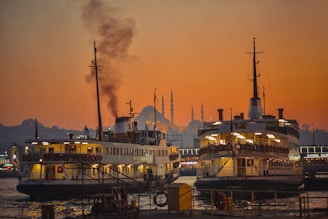 Two large ferry boats are docked at a pier with a prominent mosque featuring multiple minarets visible in the background. The sky is illuminated with a warm orange hue as the sun sets, casting a reflective glow on the water. Smoke emerges from one of the ferry chimneys, adding to the bustling atmosphere.
