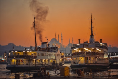 Two large ferry boats are docked at a pier with a prominent mosque featuring multiple minarets visible in the background. The sky is illuminated with a warm orange hue as the sun sets, casting a reflective glow on the water. Smoke emerges from one of the ferry chimneys, adding to the bustling atmosphere.