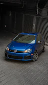 A blue Volkswagen car is parked in a dimly lit garage with a sloped concrete floor. The car is a sporty hatchback model with sleek design features, including a sunroof and alloy wheels.