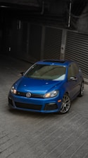 A blue Volkswagen car is parked in a dimly lit garage with a sloped concrete floor. The car is a sporty hatchback model with sleek design features, including a sunroof and alloy wheels.