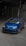 A blue Volkswagen car is parked in a dimly lit garage with a sloped concrete floor. The car is a sporty hatchback model with sleek design features, including a sunroof and alloy wheels.