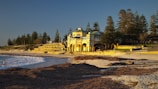 A large beachfront building with classic architecture stands prominently by the shore. The structure is surrounded by lush trees and grassy areas. The beach, with its golden sand, stretches out towards the calm blue ocean, where a person is wading in the water. The setting sun casts a warm glow over the scene.