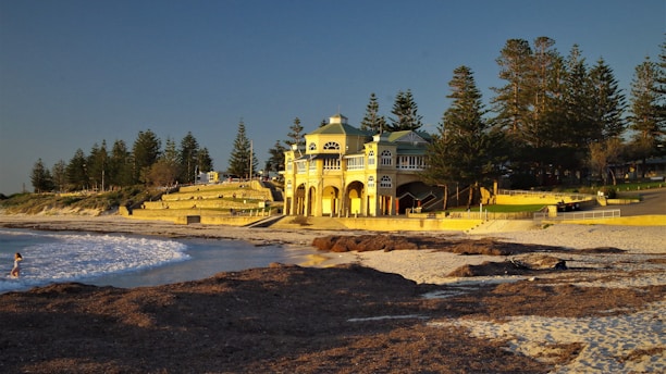 A large beachfront building with classic architecture stands prominently by the shore. The structure is surrounded by lush trees and grassy areas. The beach, with its golden sand, stretches out towards the calm blue ocean, where a person is wading in the water. The setting sun casts a warm glow over the scene.