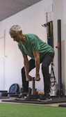 An older man is performing a strength training exercise in a gym. He is wearing a green shirt and dark pants, holding a weight with both hands. The background includes gym equipment such as weight plates and resistance bands.