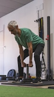 An older man is performing a strength training exercise in a gym. He is wearing a green shirt and dark pants, holding a weight with both hands. The background includes gym equipment such as weight plates and resistance bands.
