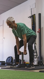 An older man is performing a strength training exercise in a gym. He is wearing a green shirt and dark pants, holding a weight with both hands. The background includes gym equipment such as weight plates and resistance bands.