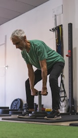 An older man is performing a strength training exercise in a gym. He is wearing a green shirt and dark pants, holding a weight with both hands. The background includes gym equipment such as weight plates and resistance bands.