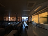 A dimly lit classroom with rows of empty desks and chairs. The sunlight is casting warm shadows across the room, illuminating the whiteboard and walls. Curtains are drawn partly, while a person appears to be sitting at the teacher’s desk near the window.