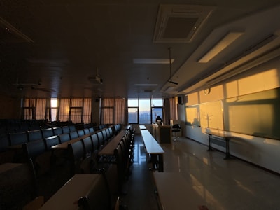 A dimly lit classroom with rows of empty desks and chairs. The sunlight is casting warm shadows across the room, illuminating the whiteboard and walls. Curtains are drawn partly, while a person appears to be sitting at the teacher’s desk near the window.