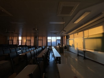 A dimly lit classroom with rows of empty desks and chairs. The sunlight is casting warm shadows across the room, illuminating the whiteboard and walls. Curtains are drawn partly, while a person appears to be sitting at the teacher&rsquo;s desk near the window.