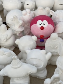 A group of sculpted figurines are gathered together, mostly in white with one prominent red and pink figure standing out. The figures appear to be cartoonish characters with varying shapes, including round and animal-like forms. They are intricately designed with different facial expressions and accessories.