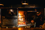A chef is working near a wood-fired oven with glowing flames. The setting has a warm, rustic ambiance with the use of dark tones and prominent lighting from the fire. The chef is wearing a dark apron and a cap, focused on preparing food.