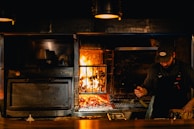 A chef is working near a wood-fired oven with glowing flames. The setting has a warm, rustic ambiance with the use of dark tones and prominent lighting from the fire. The chef is wearing a dark apron and a cap, focused on preparing food.