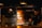 A chef is working near a wood-fired oven with glowing flames. The setting has a warm, rustic ambiance with the use of dark tones and prominent lighting from the fire. The chef is wearing a dark apron and a cap, focused on preparing food.
