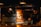 A chef is working near a wood-fired oven with glowing flames. The setting has a warm, rustic ambiance with the use of dark tones and prominent lighting from the fire. The chef is wearing a dark apron and a cap, focused on preparing food.
