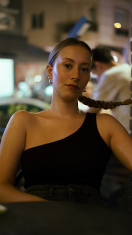 A woman with braided hair is sitting and wearing a black one-shoulder top. She appears relaxed and is looking directly at the camera. The background is softly blurred, with faint lights and a person in a white shirt visible.
