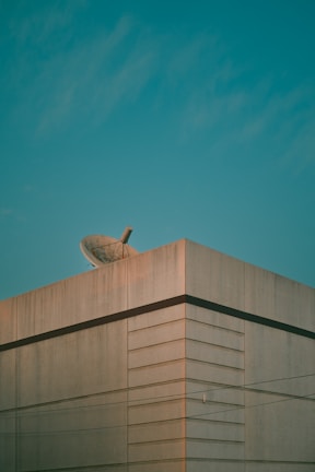 A large concrete building is topped with a satellite dish. The building's edges and lines create a geometric pattern against a clear blue sky.