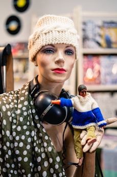 A mannequin dressed in a polka dot scarf and a knitted cap, adorned with headphones. It is holding a small figure wearing a blue and white sweater with yellow pants. The background has blurred magazine covers and vinyl records on the wall.