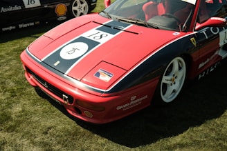 A red sports car with racing decals and the number 18 displayed prominently on its hood. The car features white rims and is parked on a grassy area. Several sponsor logos, including Shell and Girard-Perregaux, are visible on the car's body.